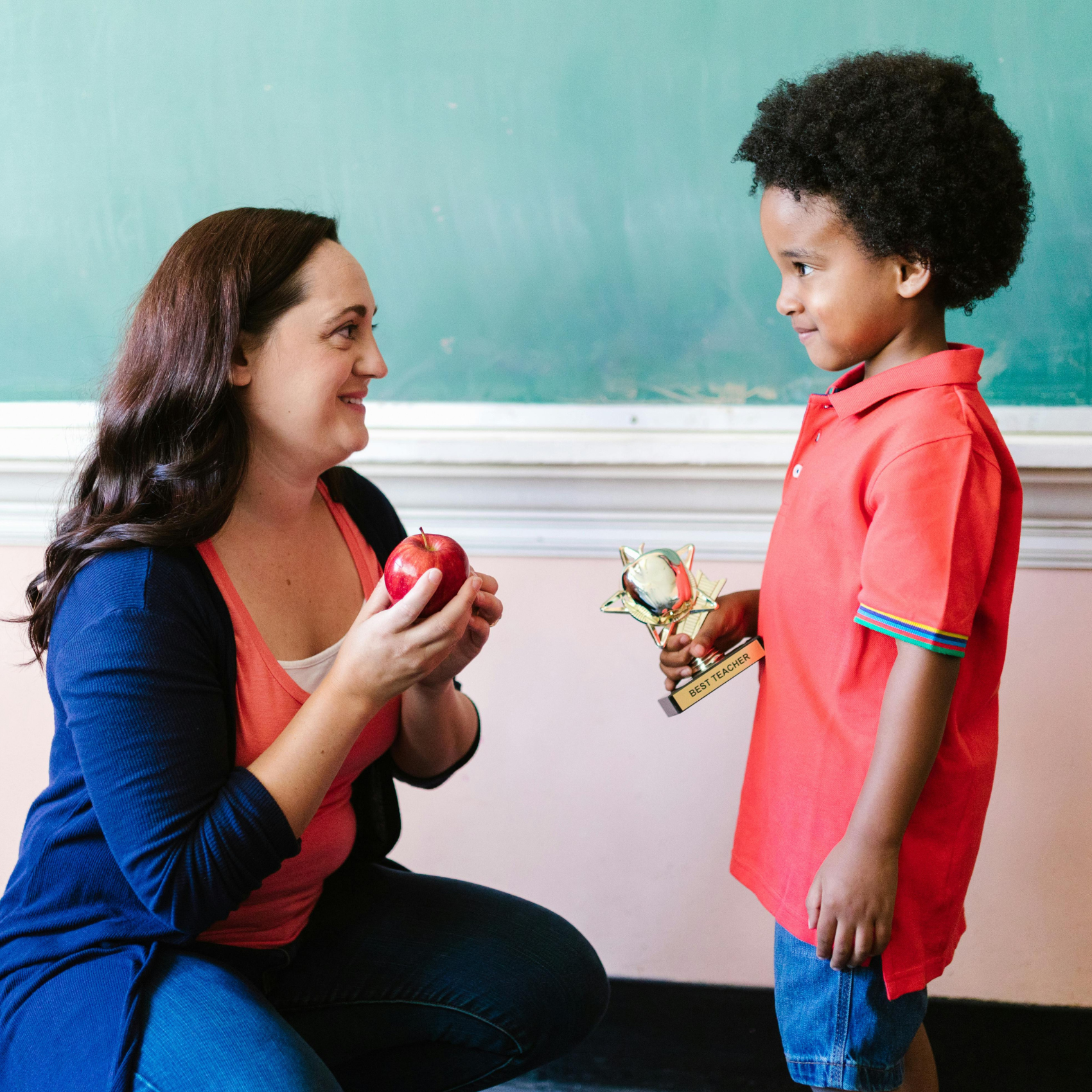 A white female teacher with long brown hair wearing a blue sweater kneels down talking to a young Black male student who is in kindergarten