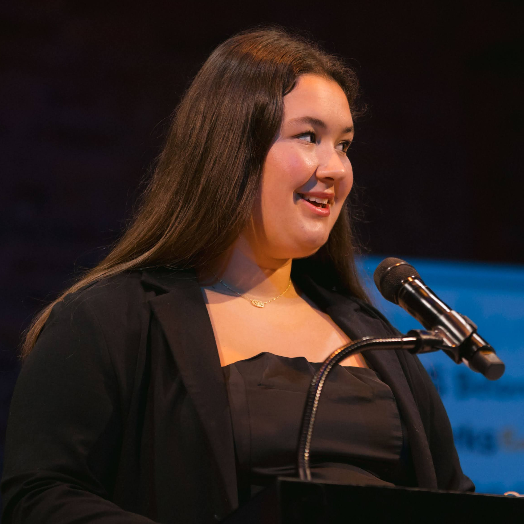 A high school girl who participants in Teens In Action speaks confidently at the podium. She is a white female with long brown hair wearing a black suit jacket.