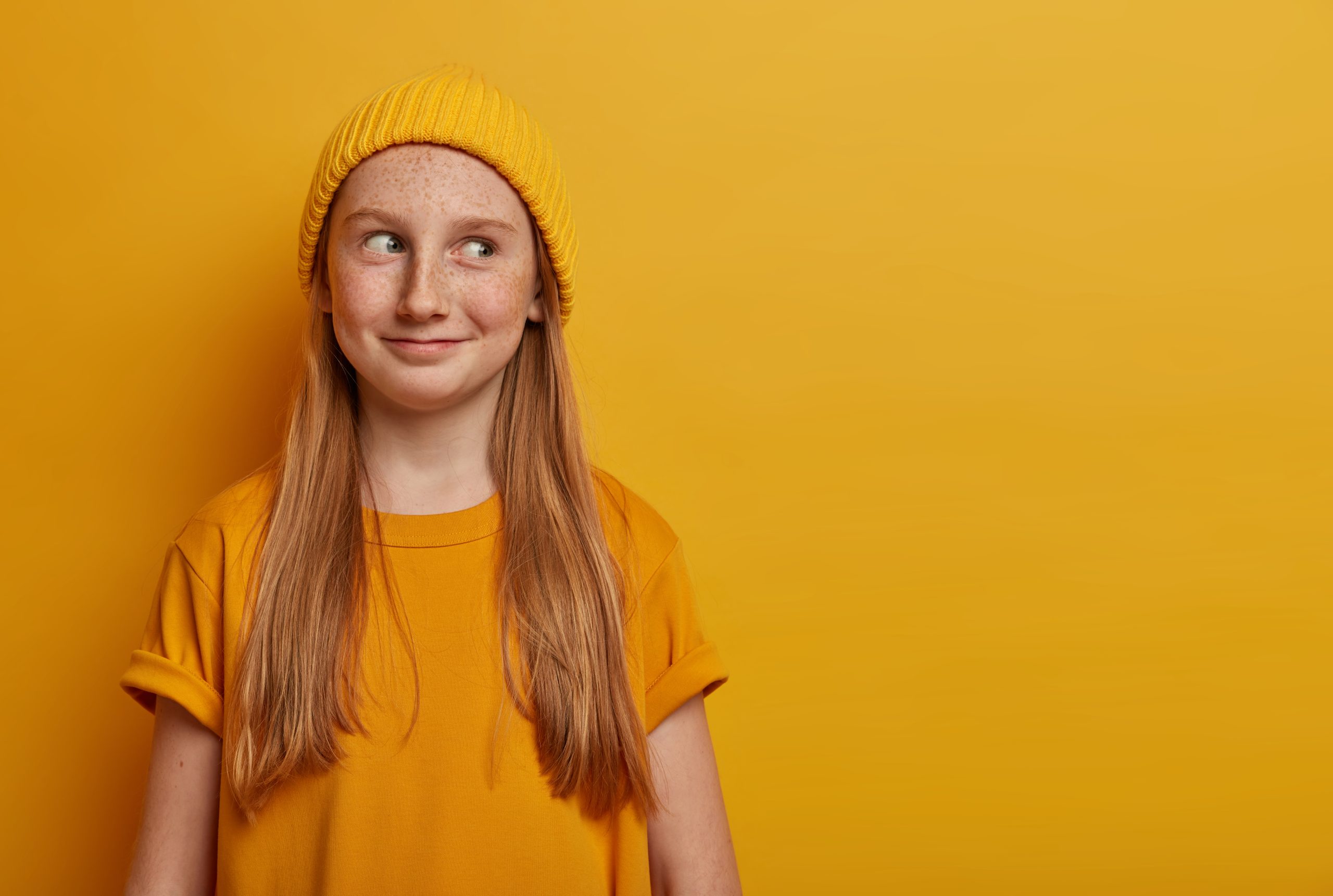 A white girl, around age 14, with strawberry blonde hair wearing a yellow hat and orange shirt against a yellow background